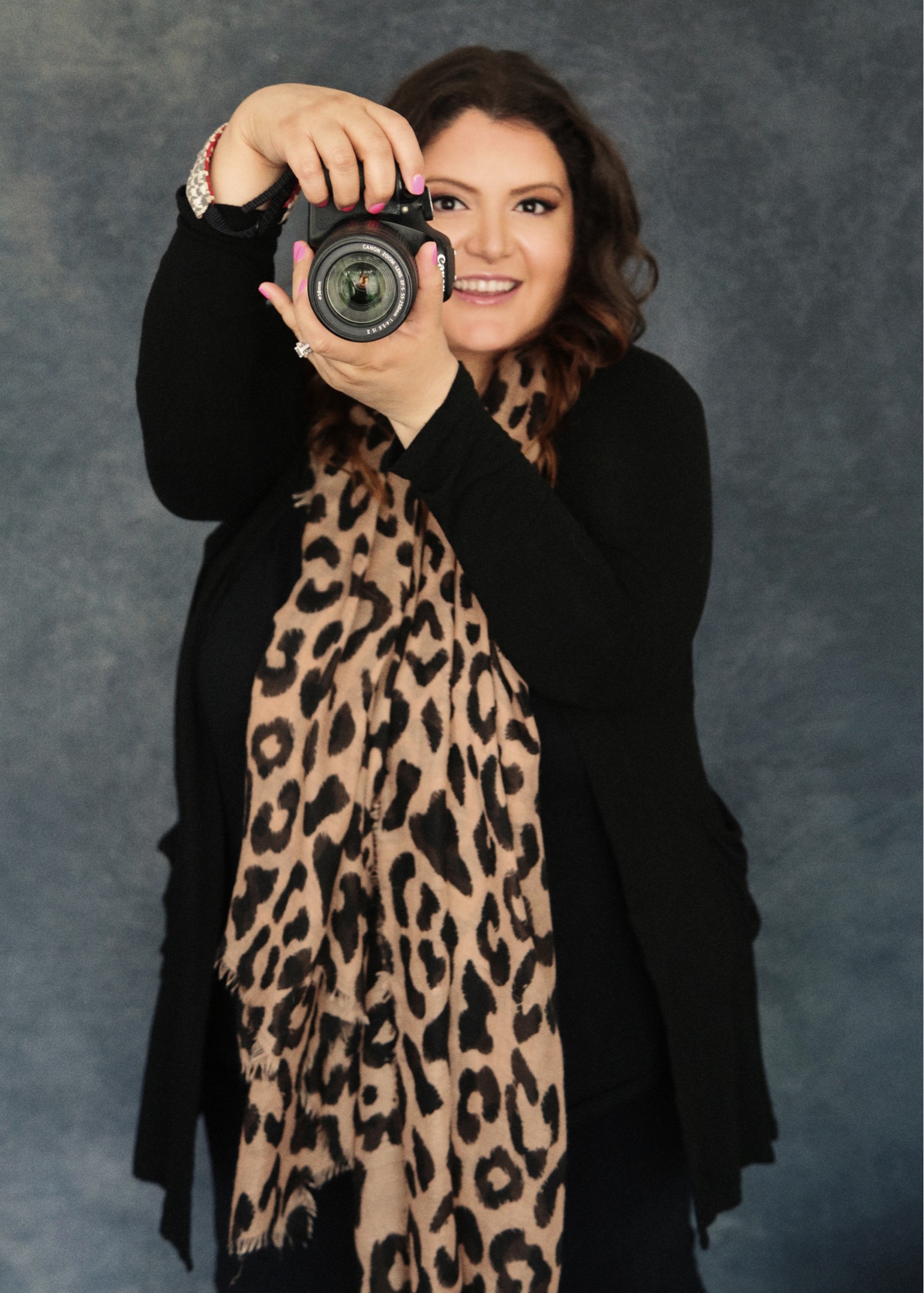 photo of Dorly Roy, photographer holding a canon camera with her pink fingernails, wearing leopard scarf. dark blue photo backdrop. she is smiling.