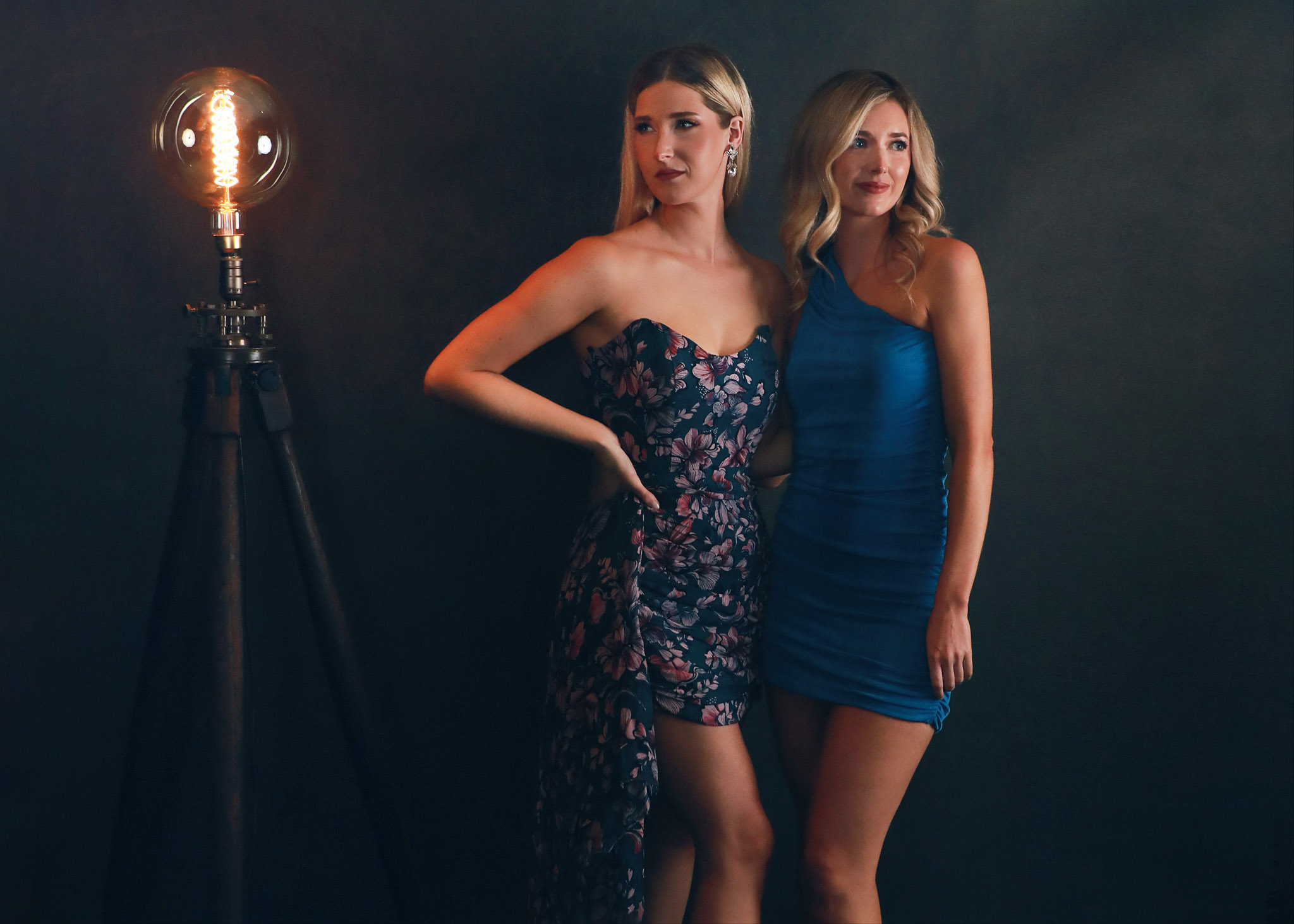 two Dallas women in ballgowns posed in a dramatic fashion next to an antique custom lamp on a dark handpainted backdrop